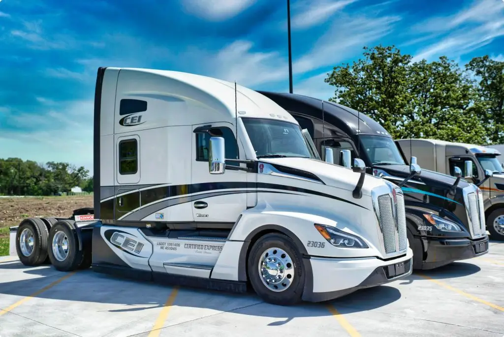 A row of 3 semi trucks are parked in three-quarter view with a blue sky and trees behind them.