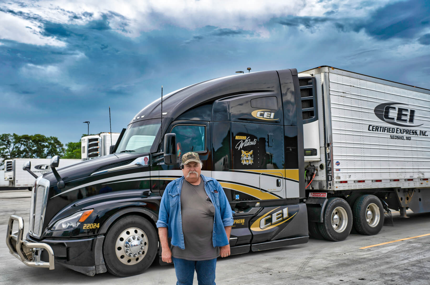 A CEI trucker stands in front of a black semi truck and trailer.