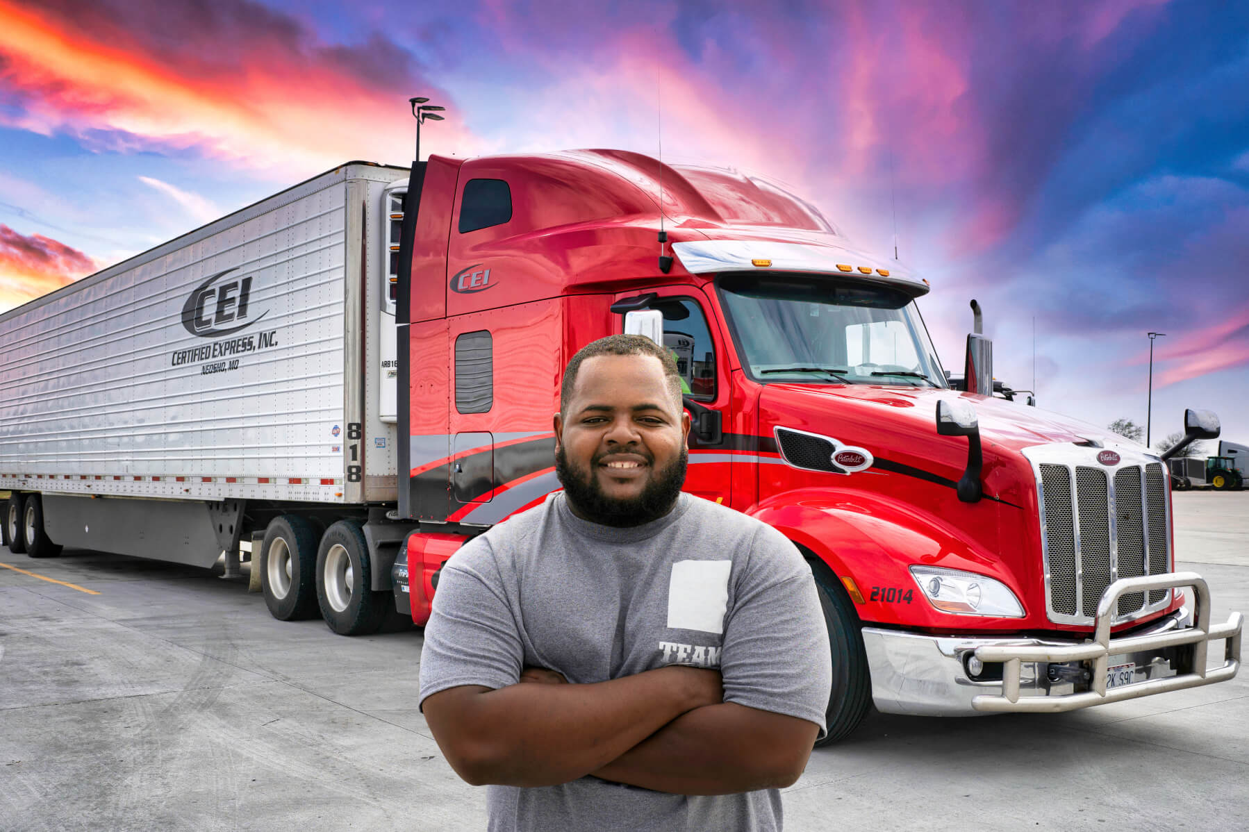 A CEI truck driver stands, arms crossed in front of a red semi truck and trailer with a colorful sunset behind.