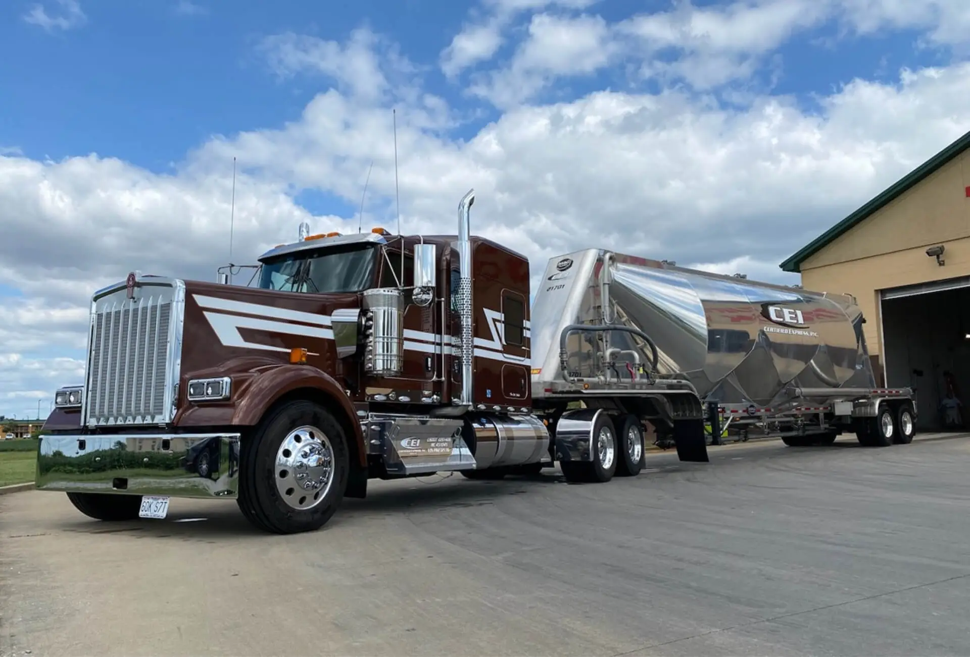 A maroon semi truck with a pneumatic trailer is parked in front of a blue sky and clouds