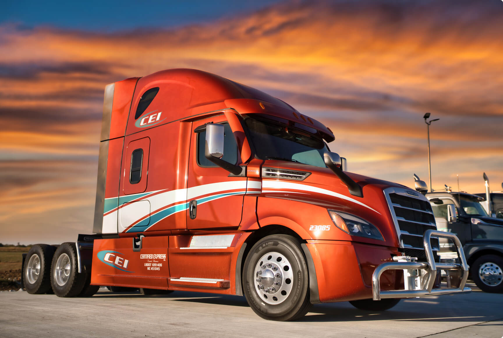A red-orange semi truck is parked beside another truck with a cloudy sunset in the background.