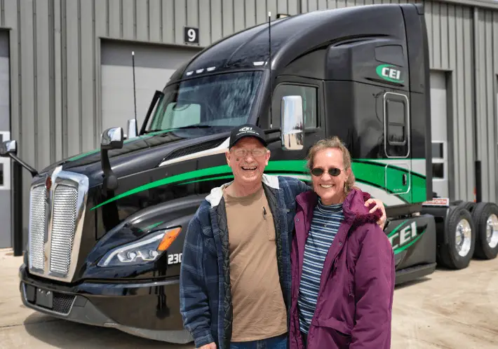 A couple with their arms around each other stands in front of a black CEI semi truck parked in front of a building.