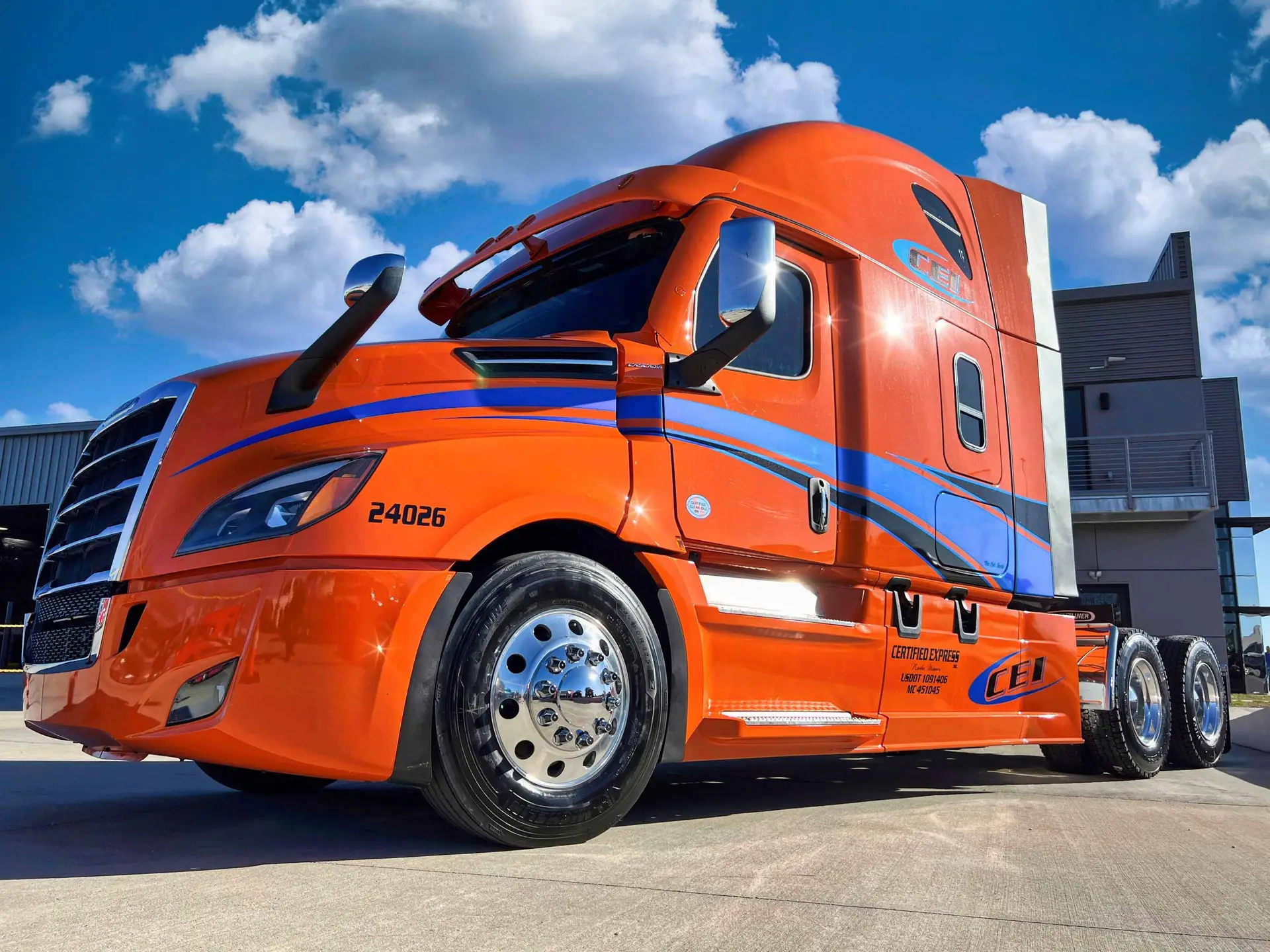 Bright orange semi-truck with blue accents parked on concrete under a blue sky with scattered clouds.