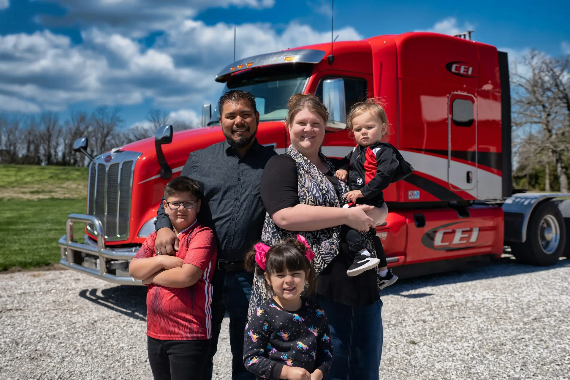 A family of five poses in front of a red CEI semi-truck on a gravel driveway under a partly cloudy sky.