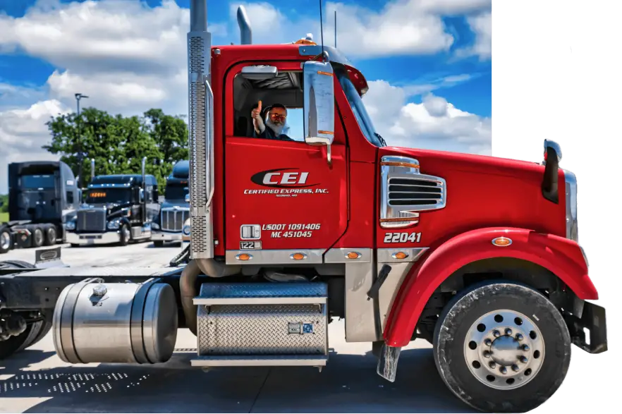 A person sits in the driver’s seat of a red CEI semi-truck, waving out the window, with several other trucks parked in the background under a partly cloudy sky.