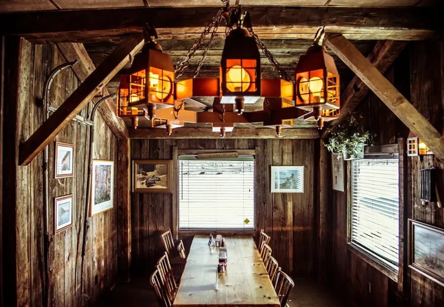 A rustic wooden dining room with a long table, wooden chairs, framed pictures on the walls, and a large chandelier hanging from the ceiling.