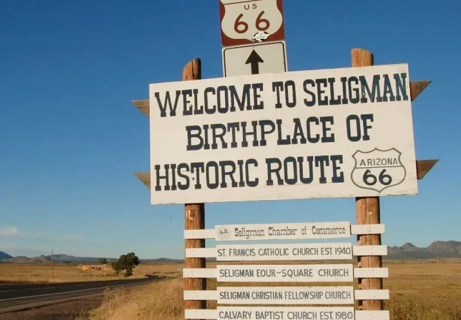 Large roadside sign reads "Welcome to Seligman Birthplace of Historic Route 66" with a list of local churches, set in a rural landscape.